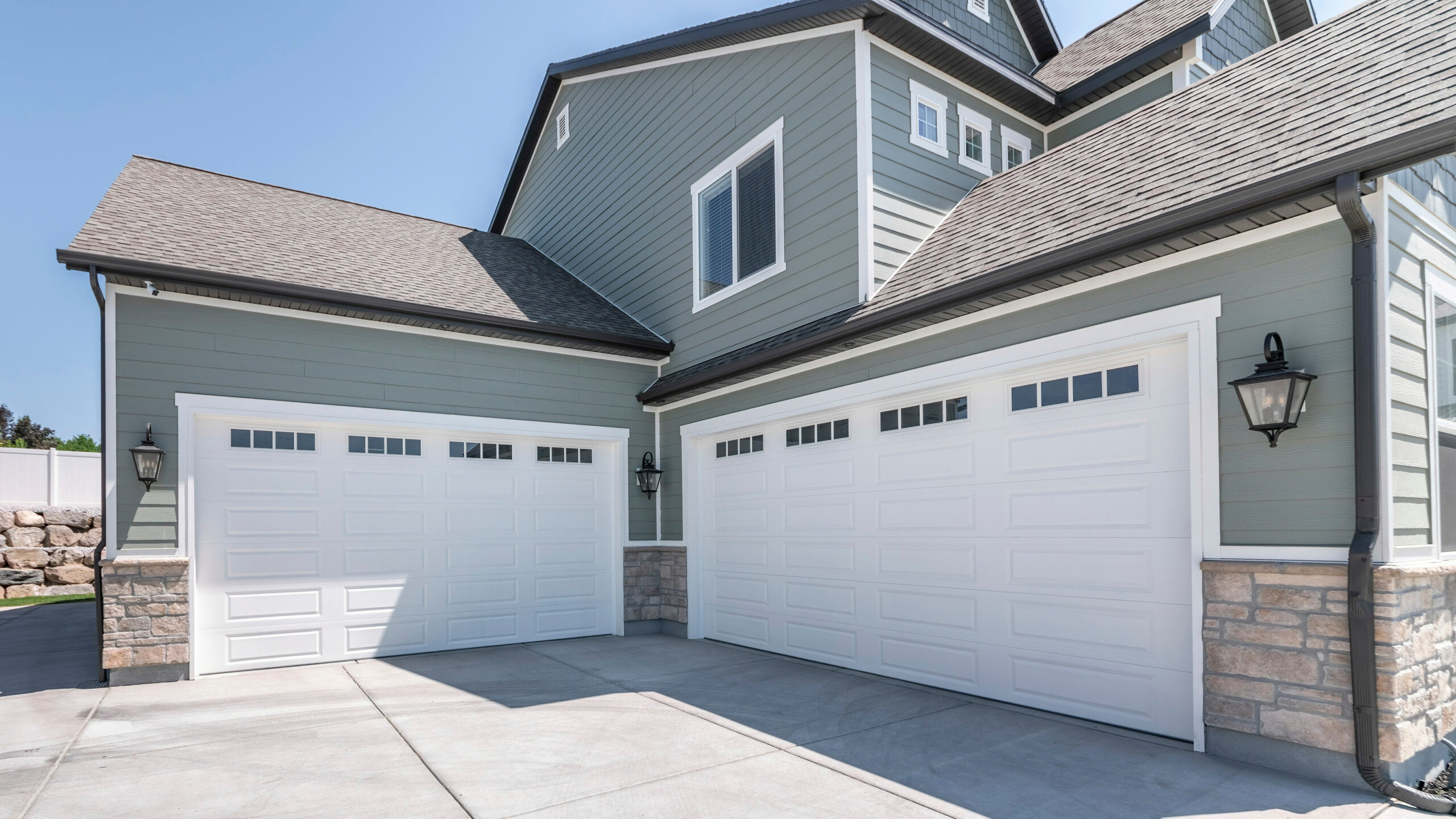 Pano Exterior of a house with concrete driveway and two closed white garage doors with windows. Side view of a gray large house with bricks and rock landscape and fence on the side.